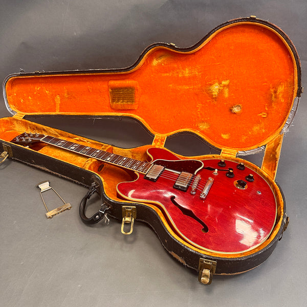 Vintage red semi-hollow electric guitar with dual humbucker pickups and tortoiseshell pickguard in an open worn black hard case with orange interior lining and detached metal bridge tailpiece