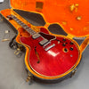 Vintage red semi-hollow electric guitar with dual humbucker pickups in an open orange plush-lined hard case with metal latches and a detached tailpiece on gray background