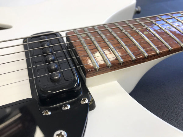 Close-up of white electric guitar showing black pickup, six steel strings, rosewood fretboard with metal frets and dot inlays, and part of the guitar body.