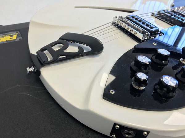 Close-up of white electric guitar body with black pickguard, chrome bridge, black control knobs labeled treble, tone, and volume, and a unique black tailpiece on a black textured surface.