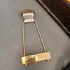 Vintage metal guitar tailpiece with diamond-shaped raised design on a gray surface next to a brown guitar case edge