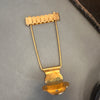 Vintage gold trapeze style guitar tailpiece with adjustable saddle pieces on a gray surface next to a brown textured item.