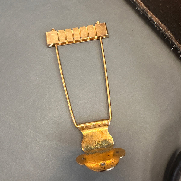 Vintage gold trapeze style guitar tailpiece with adjustable saddle pieces on a gray surface next to a brown textured item.