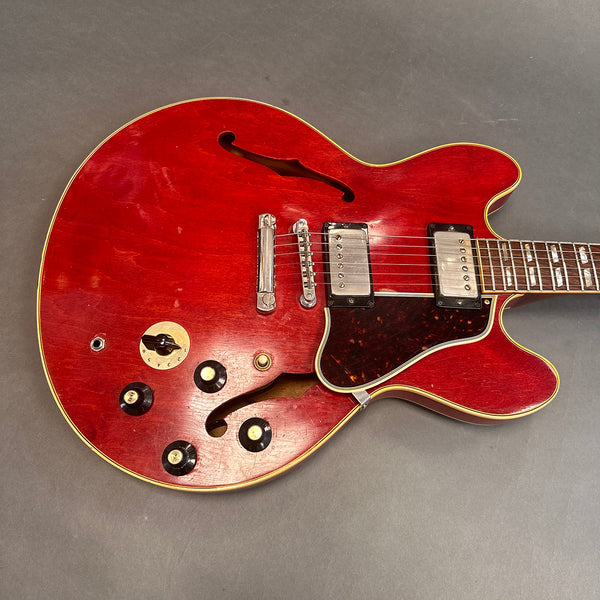 Close-up of vintage red semi-hollow body electric guitar with dual humbucker pickups, tortoiseshell pickguard, four black control knobs, and f-hole design, displayed on gray background