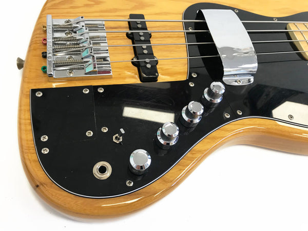 Close-up of natural wood electric bass guitar body with chrome bridge, four strings, black pickguard, control knobs, and input jack on white background
