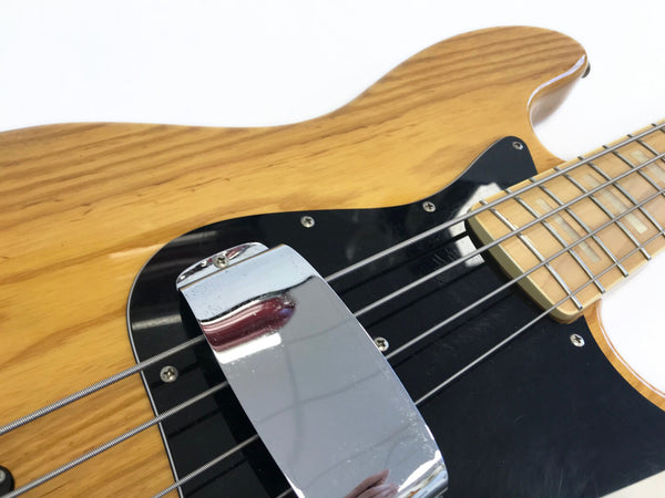 Close-up of natural wood electric bass guitar body with black pickguard, chrome pickup cover, and maple neck with block inlays