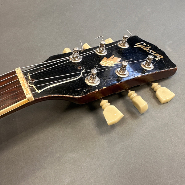 Close-up of vintage Gibson guitar headstock with tuning pegs and worn black finish, showcasing logo and tuning keys.