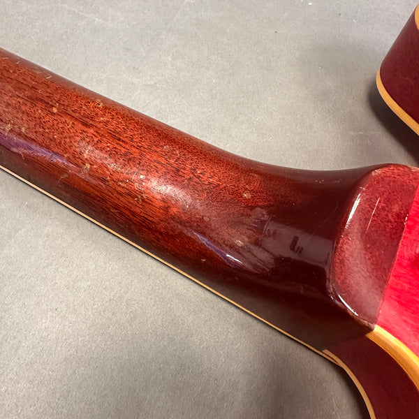 Close-up of the worn back neck and heel of a vintage guitar with reddish-brown wood finish and cream binding on a grey surface