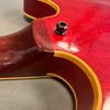 Close-up of red semi-hollow electric guitar body showing neck joint, yellow binding, and strap button on a gray surface.