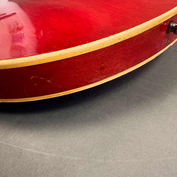 Close-up of red vintage guitar body side with cream binding and black strap button on gray surface