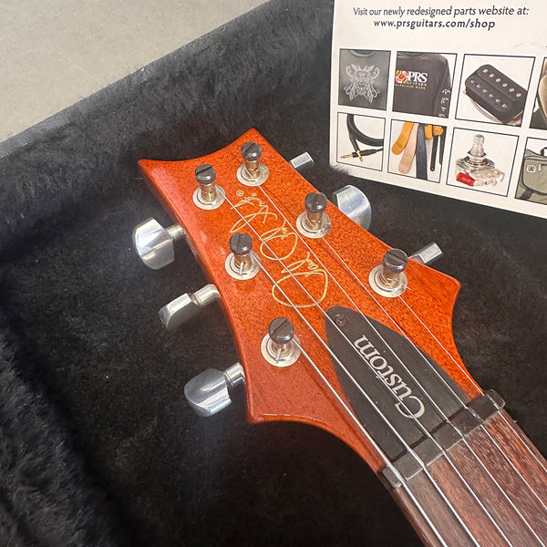 Close-up of PRS Custom guitar headstock with six tuning pegs, signature logo, and "Custom" truss rod cover on a dark plush interior guitar case