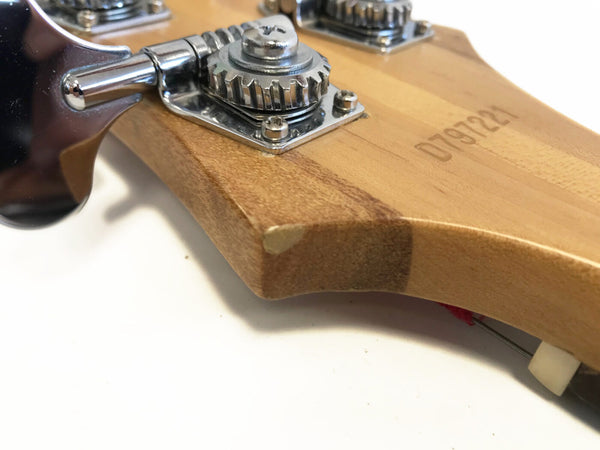 Close-up of guitar headstock corner showing tuning machine, wood grain, and serial number 0797221 with slight chip on edge.