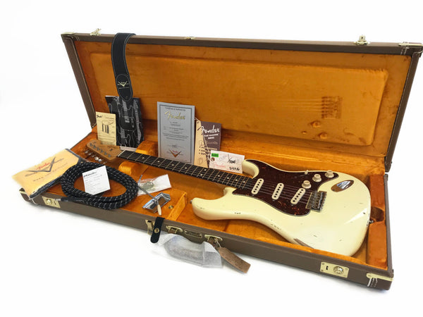 Vintage Fender Stratocaster electric guitar in white with tortoiseshell pickguard, displayed in open brown hard case with orange plush interior, including certificate of authenticity, Fender branded guitar strap, amplifier cable, and various Fender accessories and documentation.