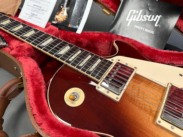 Close-up of Gibson electric guitar with sunburst finish and cream pickguard in a red plush-lined guitar case with Gibson owner's manual and inspection certificate visible