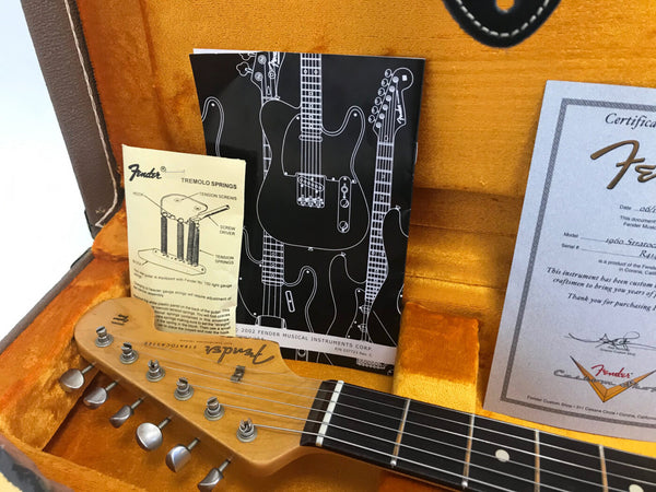 Close-up of Fender Stratocaster guitar headstock inside vintage orange plush-lined hard case with Fender tremolo springs instruction sheet, Fender guitar catalog, and official Fender certificate of authenticity partially visible.
