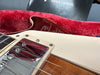 Close-up of electric guitar strings, chrome pickup, cream pickguard, and glossy wooden body in red plush-lined guitar case with owner's manual in background