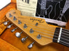 Close-up of Fender Stratocaster guitar headstock with tuning pegs inside an orange guitar case, vintage Fender instruction sheet in the background