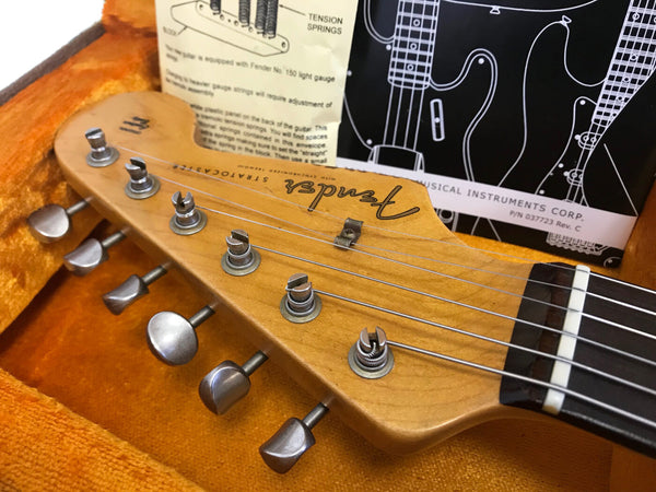 Close-up of Fender Stratocaster guitar headstock with tuning pegs inside an orange guitar case, vintage Fender instruction sheet in the background