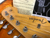 Close-up of vintage Fender Stratocaster guitar headstock with tuning pegs and string tree, showing authentic wear and original logo on maple wood, resting in orange plush guitar case with Fender maintenance instructions in background.