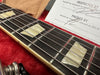 Close-up of guitar fretboard with pearloid block inlays, metal frets, and wound steel strings on a dark wood neck, resting in a red plush case with inspection and packing certificate visible.