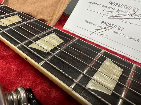 Close-up of guitar fretboard with pearloid block inlays, metal frets, and wound steel strings on a dark wood neck, resting in a red plush case with inspection and packing certificate visible.