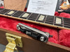 Close-up of Gibson guitar neck with pearl inlays, tremolo arm resting on red plush case lining, and inspection and packing paperwork inside Gibson guitar case
