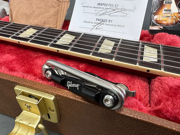 Close-up of Gibson guitar neck with pearl inlays, tremolo arm resting on red plush case lining, and inspection and packing paperwork inside Gibson guitar case