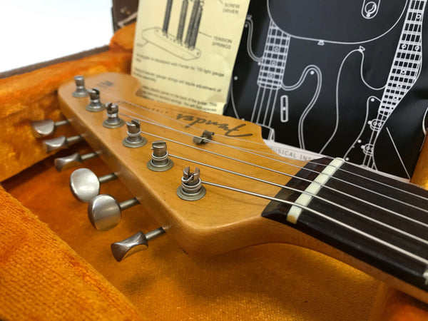 Close-up of Fender electric guitar headstock with tuning pegs, strings, and nut in orange plush guitar case with instructional sheet and guitar schematic background