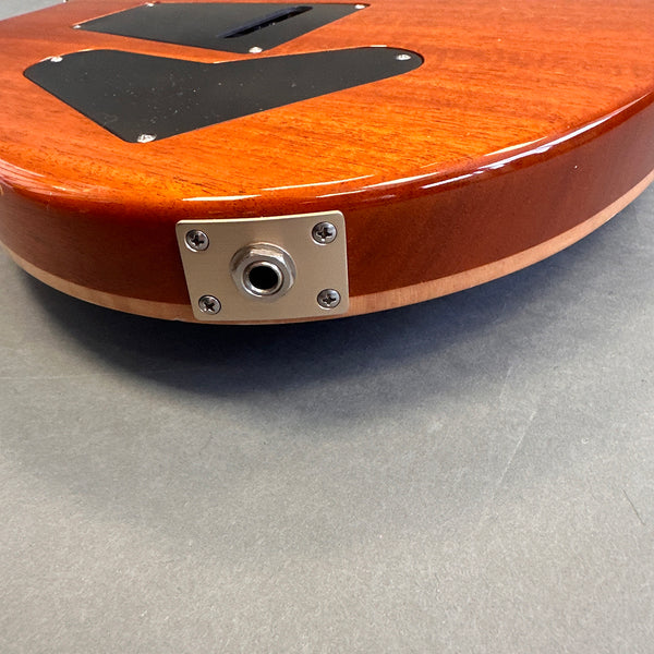 Close-up of electric guitar output jack plate on honey brown wood finish body with black backplates, displayed on grey surface.