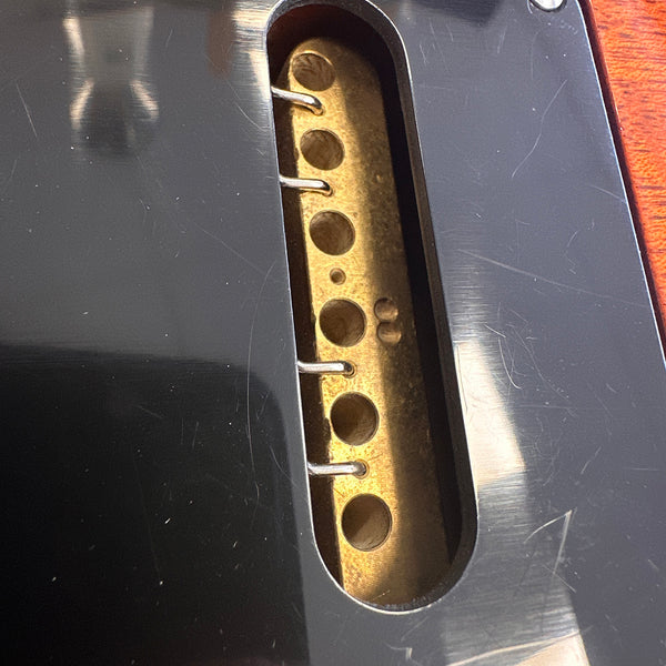 Close-up of vintage electric guitar pickup cavity with exposed brass pickup baseplate and pole pieces, surrounded by black pickguard and reddish-brown guitar body finish.