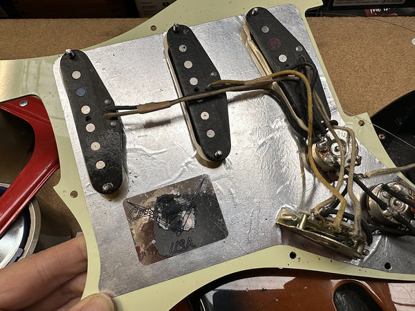 Close-up of vintage guitar pickguard showing three single-coil pickups and wiring with metal shielding and a sticker reading "Custom Shop USA"