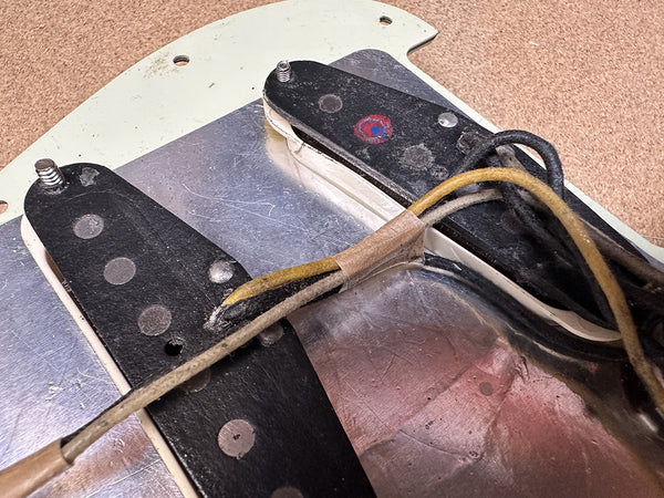Close-up of vintage guitar pickups and wiring mounted on metal and cream pickguard, showing worn black pickup covers and fabric-covered wires.