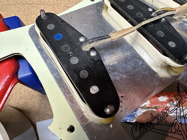 Close-up of vintage-style single coil guitar pickups mounted on metal plate with cloth-covered wiring and a portion of a pickguard showing Custom Shop lettering