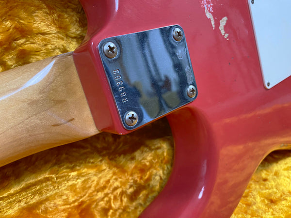 Close-up of red electric guitar neck joint with metal neck plate engraved R86958 and four screws, resting on yellow plush material