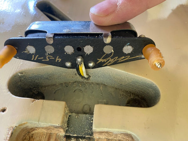 Close-up of vintage single-coil guitar pickup being held above the pickup cavity on an old cream-colored electric guitar body, showing wiring and date markings.