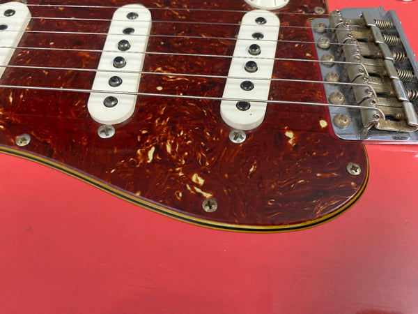 Close-up of electric guitar body showing tortoiseshell pickguard, white single-coil pickups, metal bridge, and coral red finish with visible wear and scratches