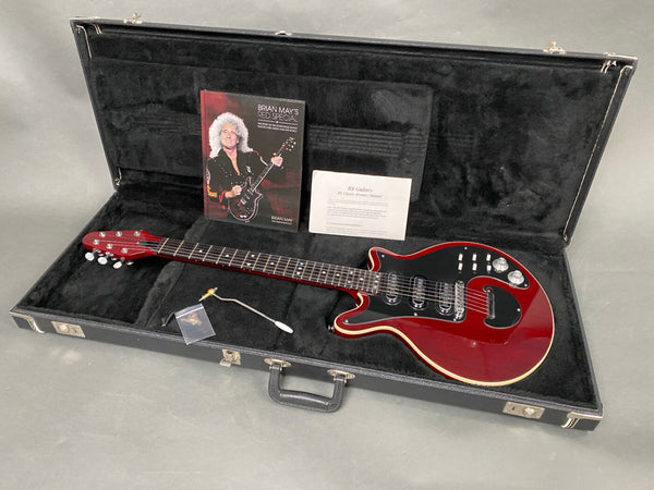 Brian May Red Special electric guitar in red with black pickguard, three pickups, and cream binding, displayed inside a black hard case with accessory compartment containing a tremolo arm, user manual, and book titled "Brian May's Red Special"