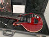 Red RS Guitars electric guitar with black pickguard, three single-coil pickups, and silver control knobs inside a black hard case with plush interior, accompanied by an RS Classic Owners Manual and a photo of Brian May playing guitar.
