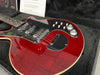 Close-up of red electric guitar with black pickguard, three chrome pickups, and silver control knobs in black plush-lined guitar case with owner's manual in background
