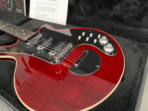 Close-up of red electric guitar with black pickguard, three chrome pickups, and silver control knobs in black plush-lined guitar case with owner's manual in background