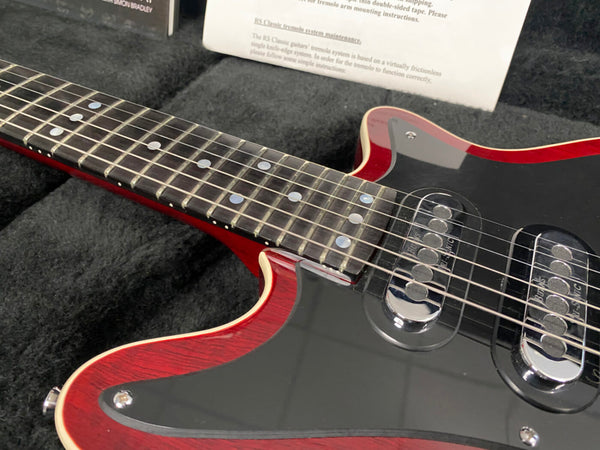Close-up of red electric guitar with black pickguard and dual humbucker pickups, neck with dot inlays, resting in black plush guitar case with documentation visible in background