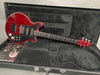 Red electric guitar with black pickguard and three pickups inside a black plush-lined hard case with brochure beneath guitar body