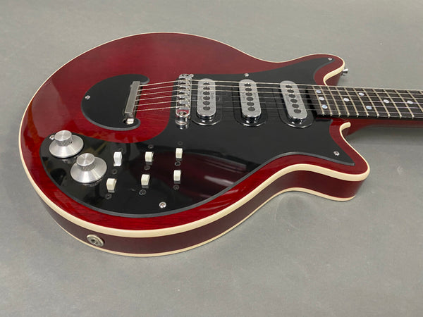Close-up of red electric guitar body with black pickguard, three single-coil pickups, two silver control knobs, and white toggle switches on gray background