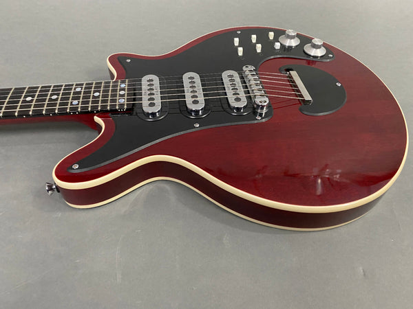 Close-up of electric guitar body with dark red finish, black pickguard, three single-coil pickups, chrome bridge, and multiple control knobs on grey background.