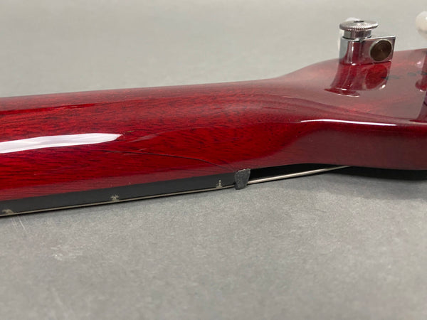 Close-up of dark red guitar neck showing cracked wood finish and metal string tuner on gray surface