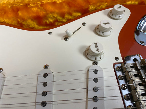 Close-up of electric guitar white pickguard with volume and tone control knobs, three single-coil pickups, and metal bridge on orange guitar body