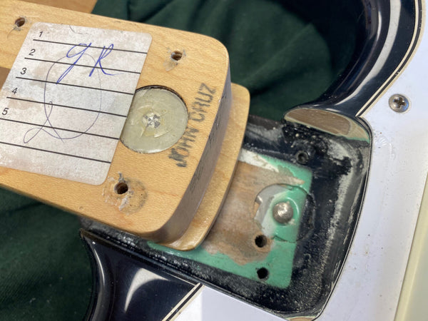 Close-up image of the neck pocket of an electric guitar showing neck heel with handwritten notes, mounting screw, and part of the black and white guitar body with visible wear and green paint inside the neck cavity.