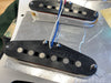 Close-up of two single-coil electric guitar pickups with black covers and pole pieces, mounted on a metal pickup plate with blue and white wires attached, shown during guitar assembly or repair.