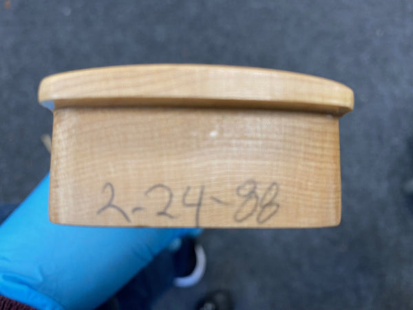 Close-up of a light wood guitar neck heel with handwritten date "2-24-98" held by a person wearing a blue glove, showing wood grain details.
