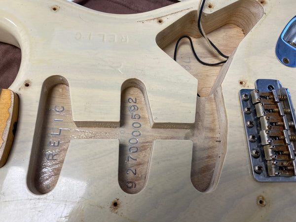 Close-up of unfinished natural wood electric guitar body showing stamped serial number 9270000592 and rider shield pickguard cavity with RELIC imprint and vintage style tremolo bridge hardware
